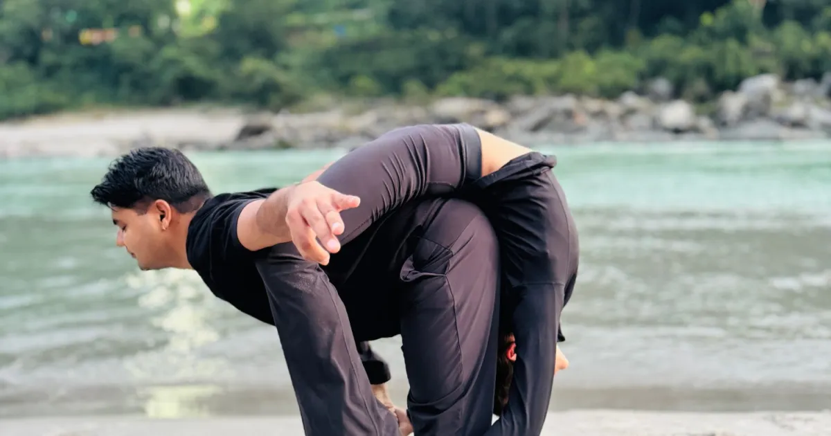 A standing acro yoga base carrying a partner across his back on the sand beside the Ganga in Rishikesh