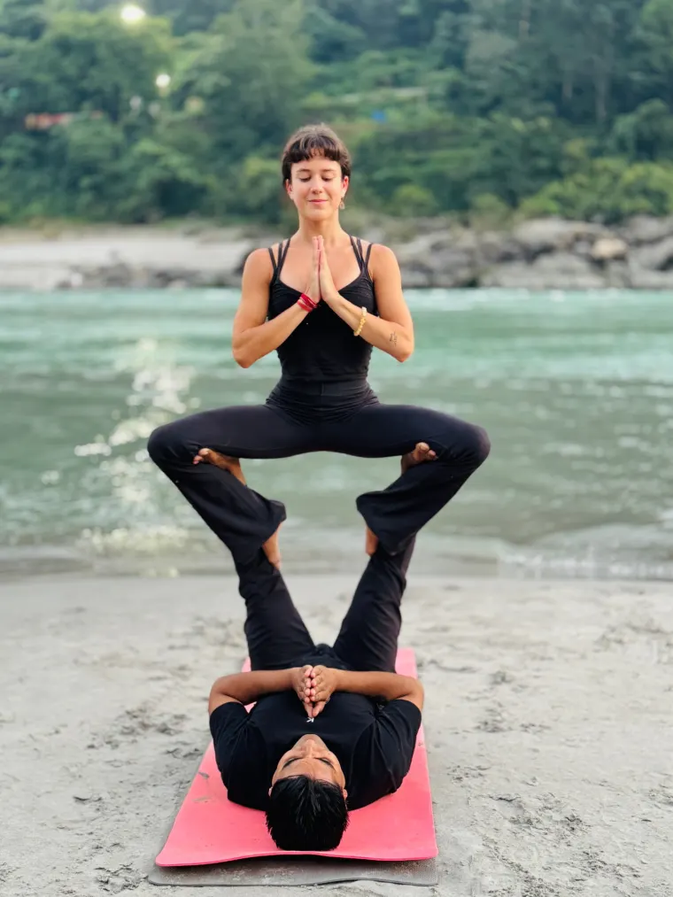 Two students practising a supported partner yoga balance on the sand beside the Ganga in Rishikesh, one balanced on the other's feet with hands in namaste