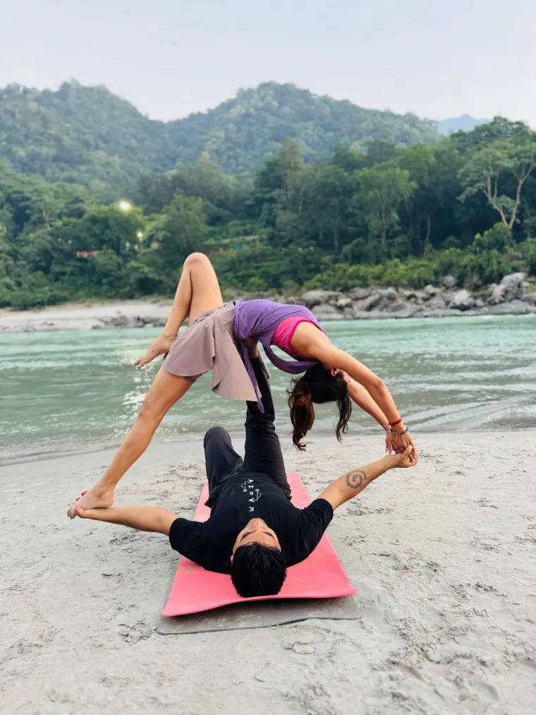 An acro yoga flyer in a supported backbend on the base's feet, practising on the sand beside the Ganga in Rishikesh